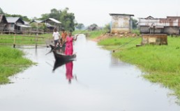 Woman in a boat in a river with village houses on the banks. Credit: NEADS